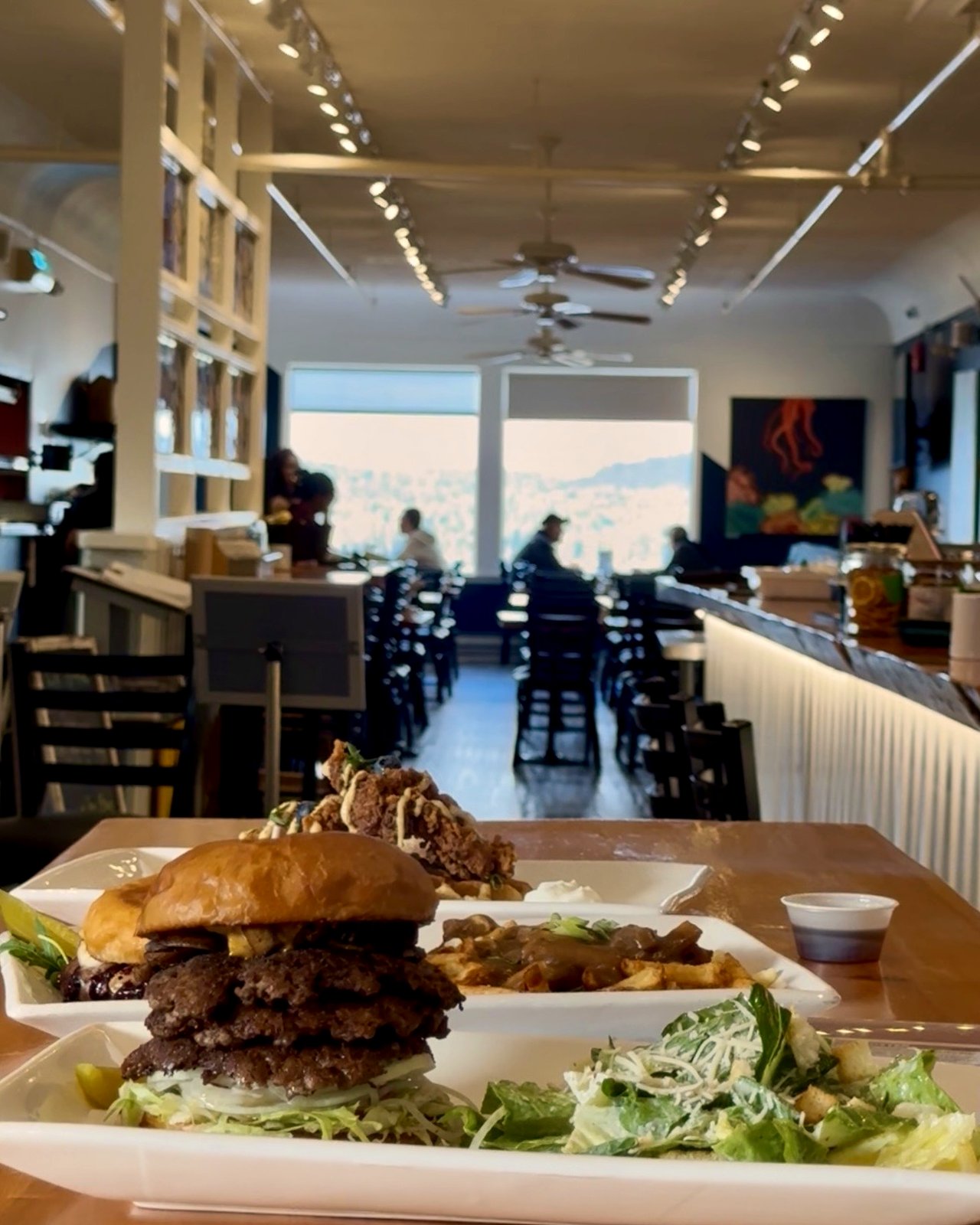 A spread of OV burgers, poutine, and Caesar salad in the foreground with the OV dining room stretching to harbour-facing windows at the back