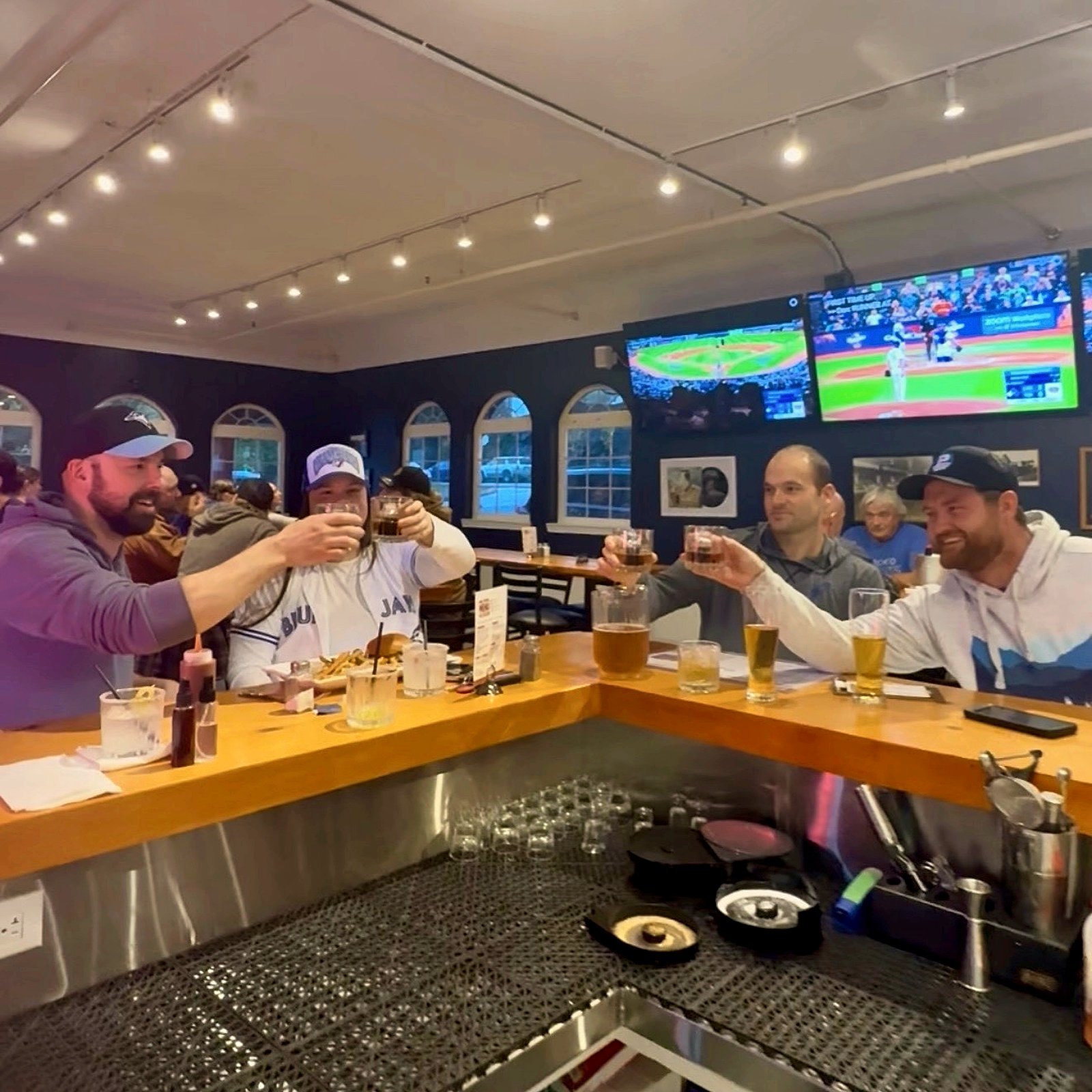 Four guests raising drinks in a toast across the bar at OV Burger Bar while a baseball game plays on the TVs, in Prince Rupert, BC